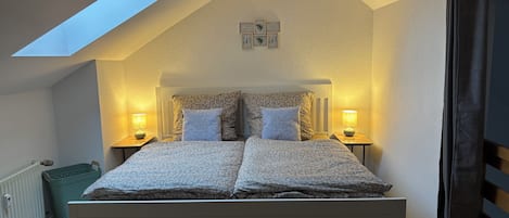 Un dormitorio con techo inclinado, tragaluz, cama con dos almohadas, dos lámparas de noche y un cuadro pequeño en la pared.