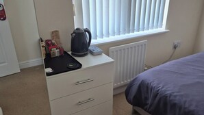 A bedroom with a bed, a radiator, a window with blinds, a desk with a kettle, and a wall-mounted sign. - Room-cozy Guest Room With Modern Ameniti (Tipton)