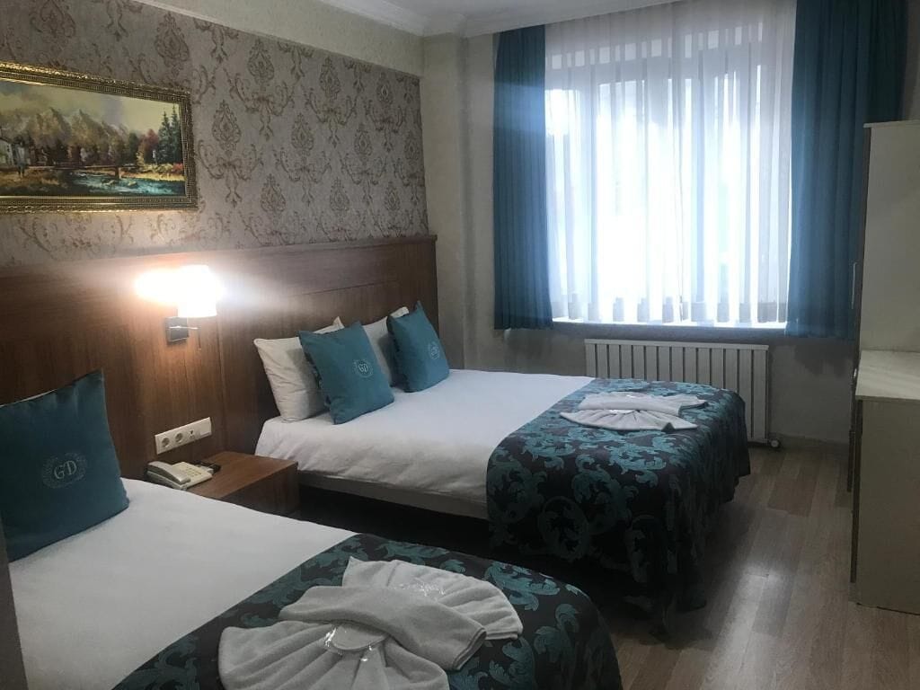 A hotel room with two beds, a painting on the wall, a window with curtains, and a radiator.