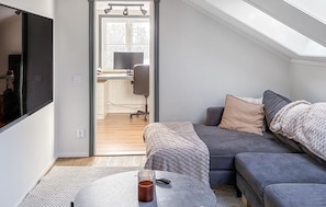 Ein modernes Wohnzimmer mit einem grauen Sofa, einem Couchtisch und einem Fernseher. Im Hintergrund steht ein Schreibtisch mit einem Computer und einem Stuhl.