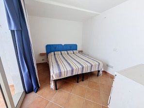 Ein kleines Zimmer mit einem Einzelbett, einem blauen Kopfteil, einer gestreiften Bettdecke und einem Fliesenboden.