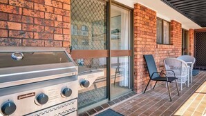 Une terrasse extérieure équipée d’un barbecue Jumbuck, une porte vitrée donnant sur un espace abrité et une chaise en osier blanche.