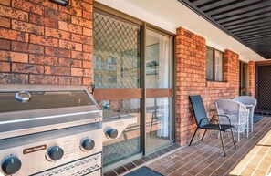 An outdoor patio with a Jumbuck barbecue grill, a glass door leading to a covered area, and a white wicker chair. - The Entrance Escape - 1 min walk to the beach (The Entrance)