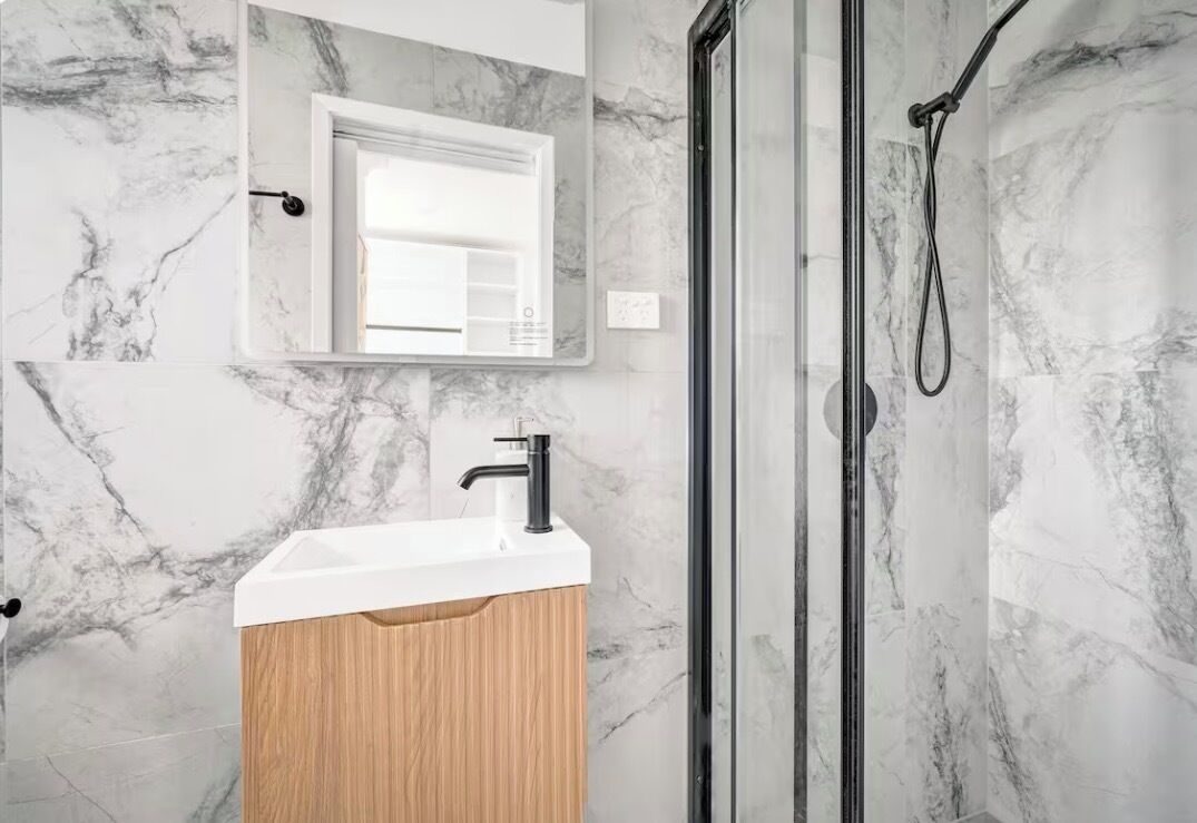 Un bagno con parete in marmo, mobile lavabo in legno, uno specchio e cabina doccia in vetro.