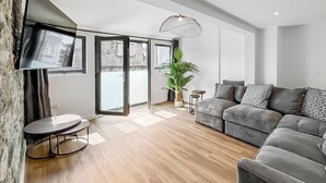 Ein modernes Wohnzimmer mit grauem Sofa, einem kleinen runden Tisch, einem Blumentopf und einem an der Wand befestigten Fernseher.