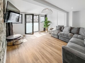 Ein modernes Wohnzimmer mit grauem Sofa, einem kleinen runden Tisch, einem Blumentopf und einem an der Wand befestigten Fernseher.