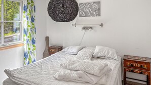 Ett sovrum med en säng, ett sängbord, en lampa, ett fönster med gardiner och ett konstverk på väggen.