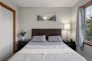 Ein Schlafzimmer mit einem Bett, einem Nachttisch, einer Wandlampe, einer Uhr, einem gerahmten Bild und einem Fenster mit Vorhängen.