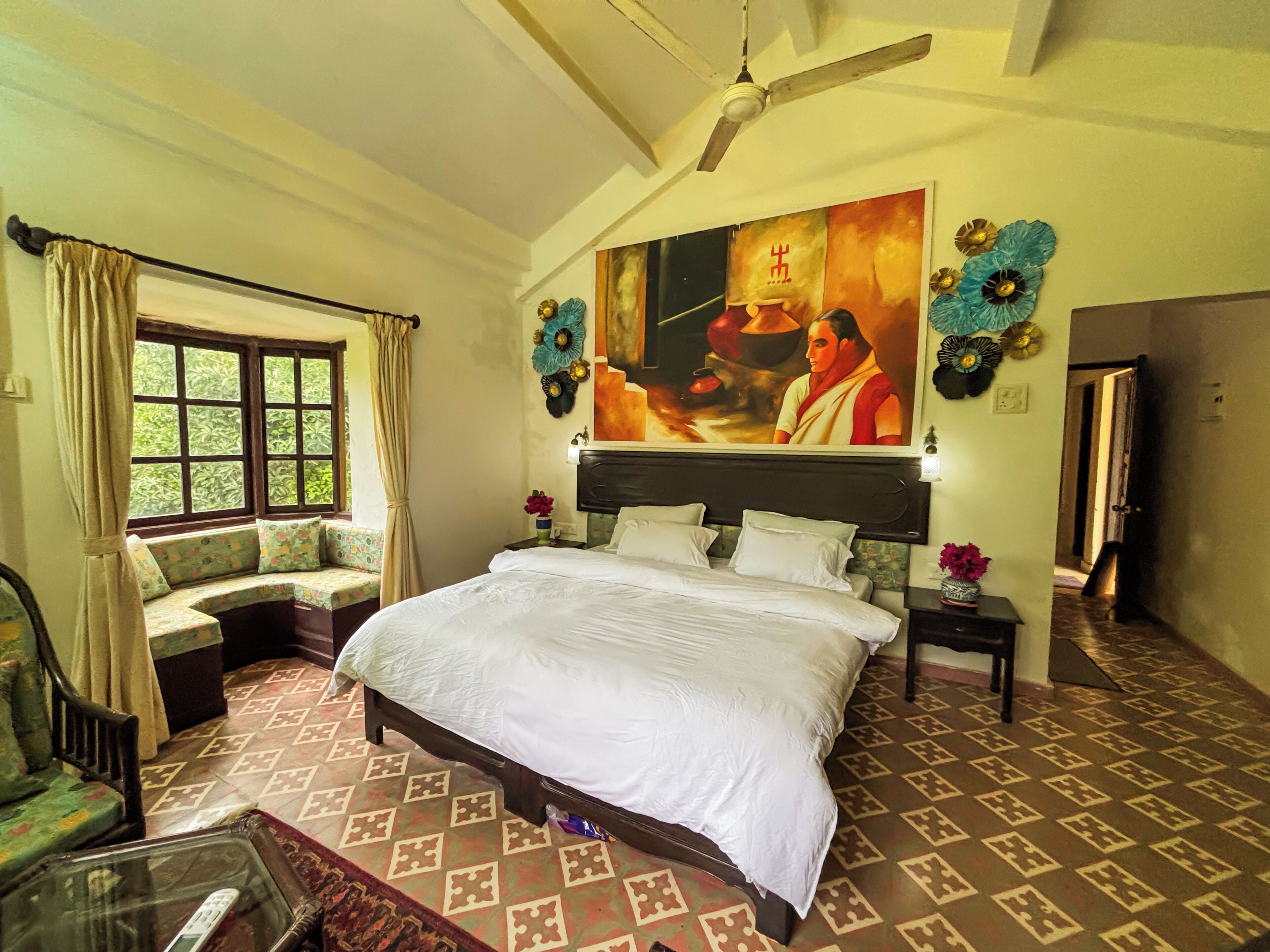 A hotel room with a large bed, a ceiling fan, a window with curtains, a bench by the window, and a wall with a painting.