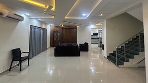 Ampla sala de estar com sofá, poltrona, escada e cozinha ao fundo.
