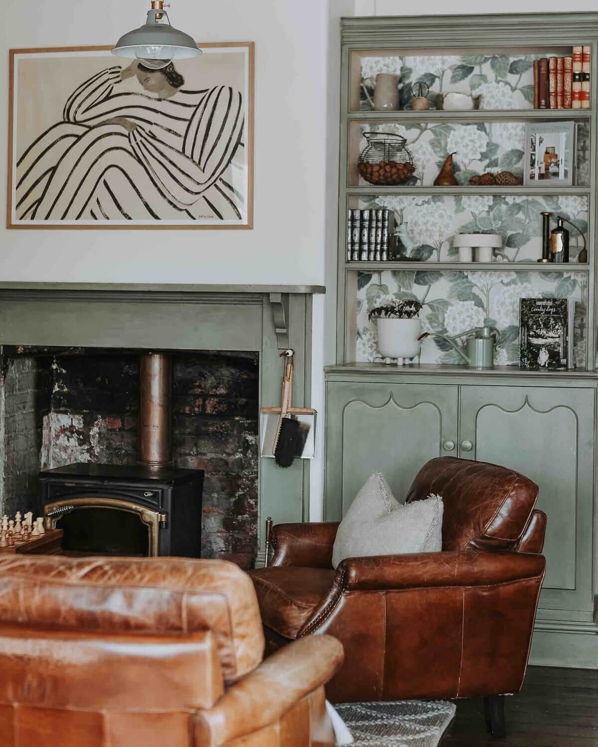Een woonkamer met een open haard, een leren fauteuil, een boekenkast met decoratieve objecten, en een lijst met een abstract schilderij.