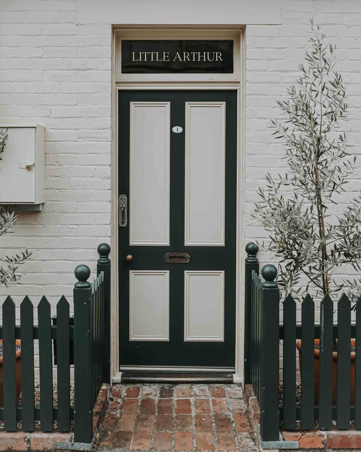 Una puerta negra con el nombre 'Little Arthur' encima, un buzón y un árbol pequeño al lado de la puerta.
