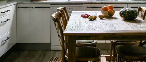 Una cocina con ventana, gabinetes blancos, una mesa de madera con sillas y una lámpara colgante.