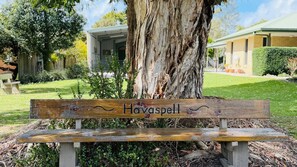 Een houten bank met een bord waarop 'Havaspell' staat, voor een grote boom.