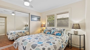 A bedroom with a bed, a bedside table, a lamp, a window with blinds, a mirror, and a door. - 5 Zircon Street - Rainbow Beach (Rainbow Beach)