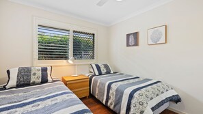 Een slaapkamer met twee bedden, een nachtkastje, een raam met jaloezieën en twee ingelijste foto's aan de muur.