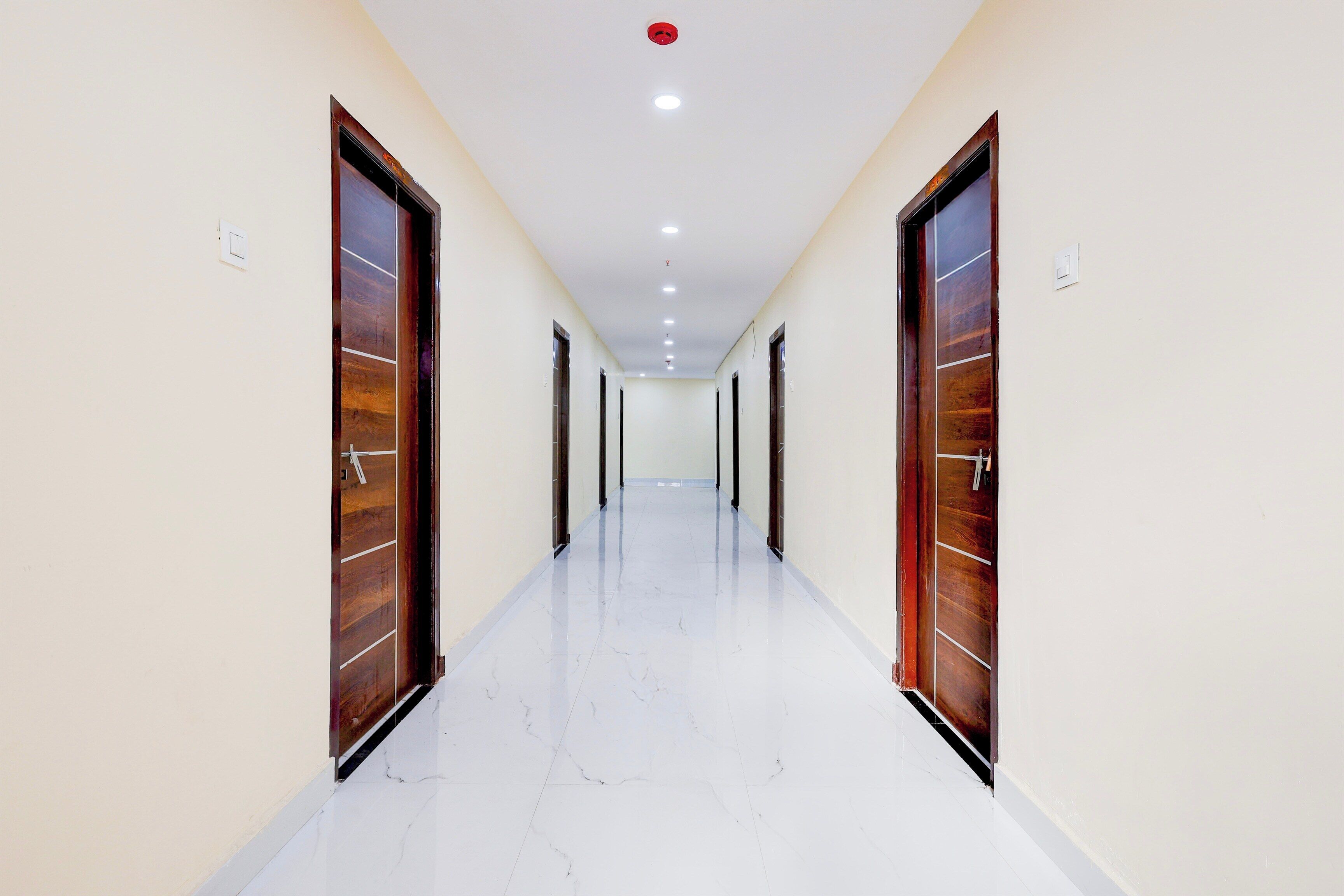 Un couloir bordé de portes en bois de chaque côté, avec un sol carrelé de carreaux blancs et des spots encastrés au plafond.