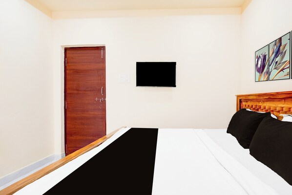 Habitación de hotel con puerta de madera, un televisor de pantalla plana montado en la pared, una cama con ropa de cama blanco y negro, y cabecero de madera.