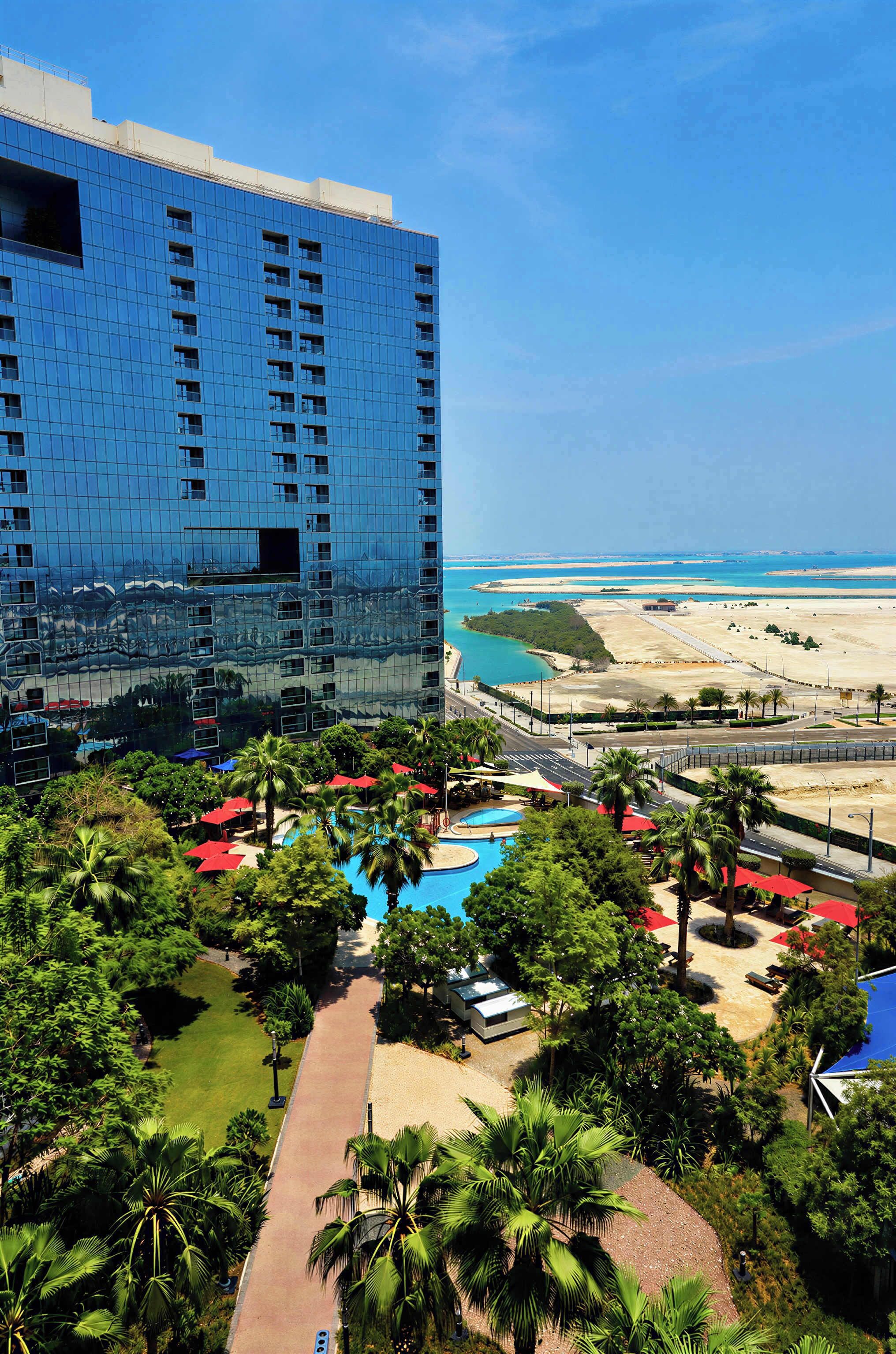 A high-rise hotel with a reflective glass facade, a pool area with palm trees, and a view of the beach and ocean.