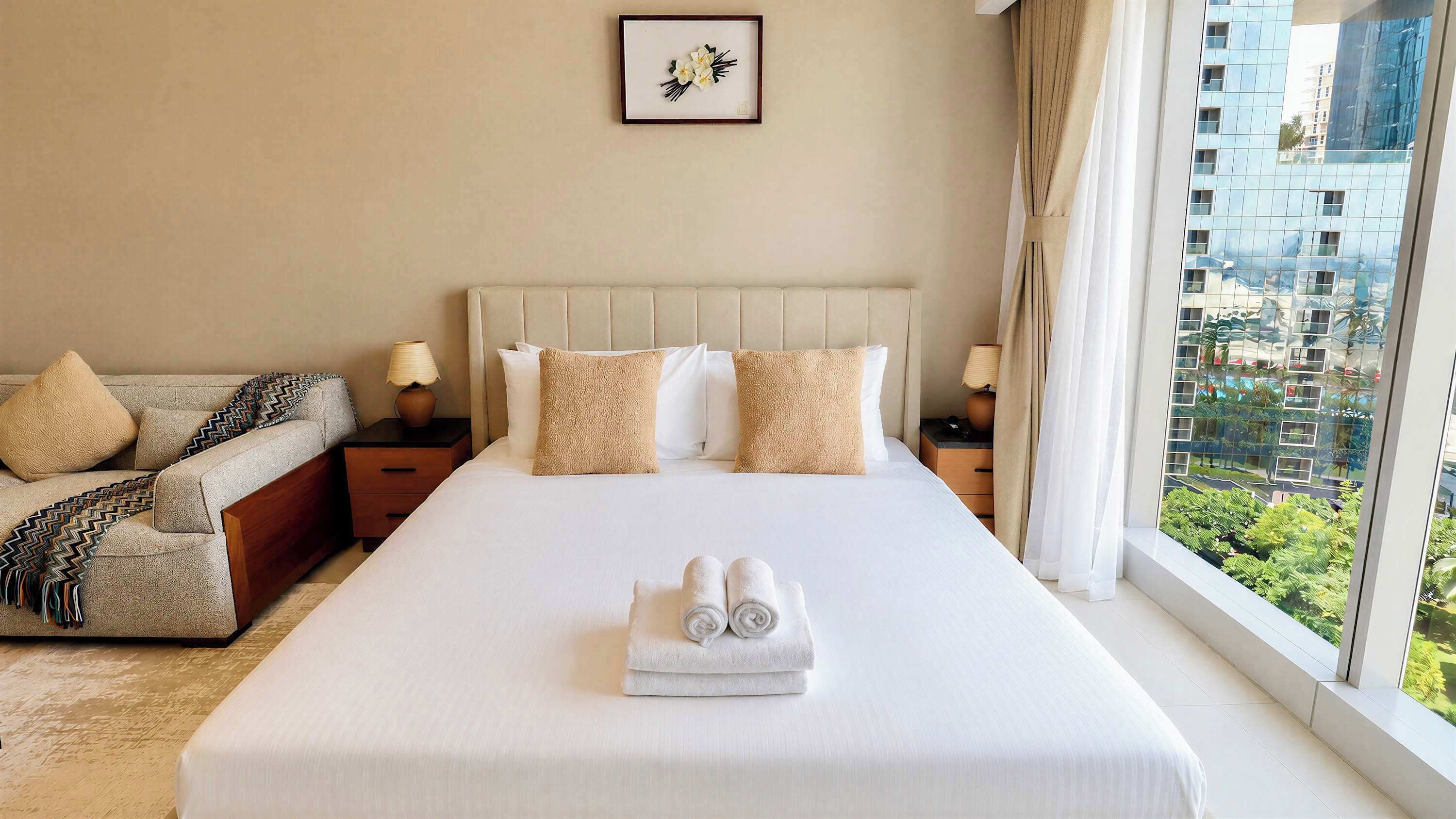 A hotel room with a large bed, two bedside tables, a sofa, and a window with a city view.