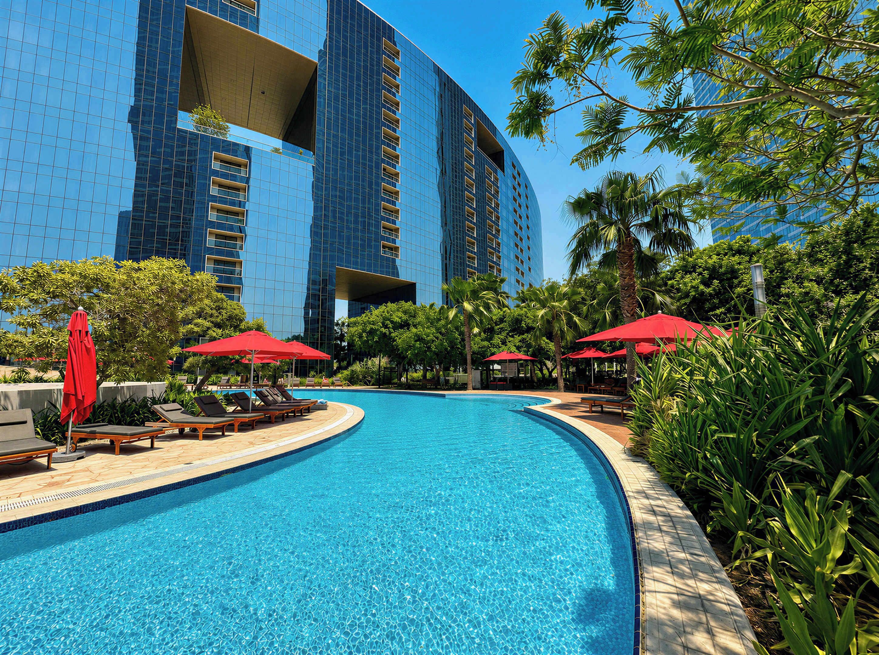 A swimming pool with lounge chairs and red umbrellas, surrounded by palm trees and a modern building.