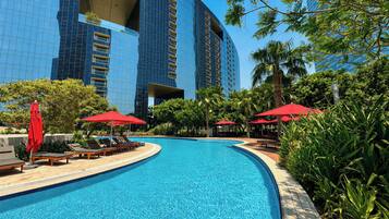 A swimming pool with lounge chairs and red umbrellas, surrounded by palm trees and a modern building.