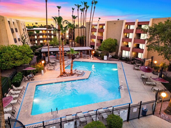 A hotel pool area with multiple lounge chairs and umbrellas, surrounded by palm trees and a multi-story building with balconies. - Resort Pool + Spa + Balcony | Fashion Square + Echo Canyon | Camelback Cabana (Scottsdale)