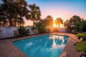 Une piscine avec vue sur le coucher de soleil, entourée de palmiers et de chaises longues.
