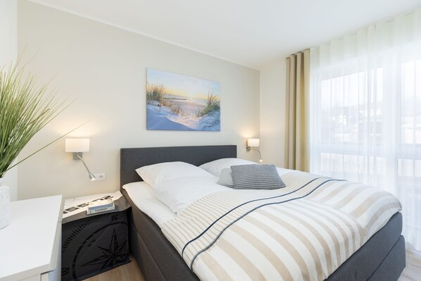 Siististi järjestetty hotellihuone, jossa on suuri sänky, yöpöytä lampun kanssa, taulu seinällä ja ikkuna, jossa on ohuet verhot.