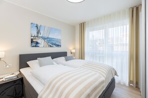 Hotellihuone, jossa on suuri sänky, kaksi yöpöytää, yölamppu, purjelaivan kuva ja läpikuultavat verhot.