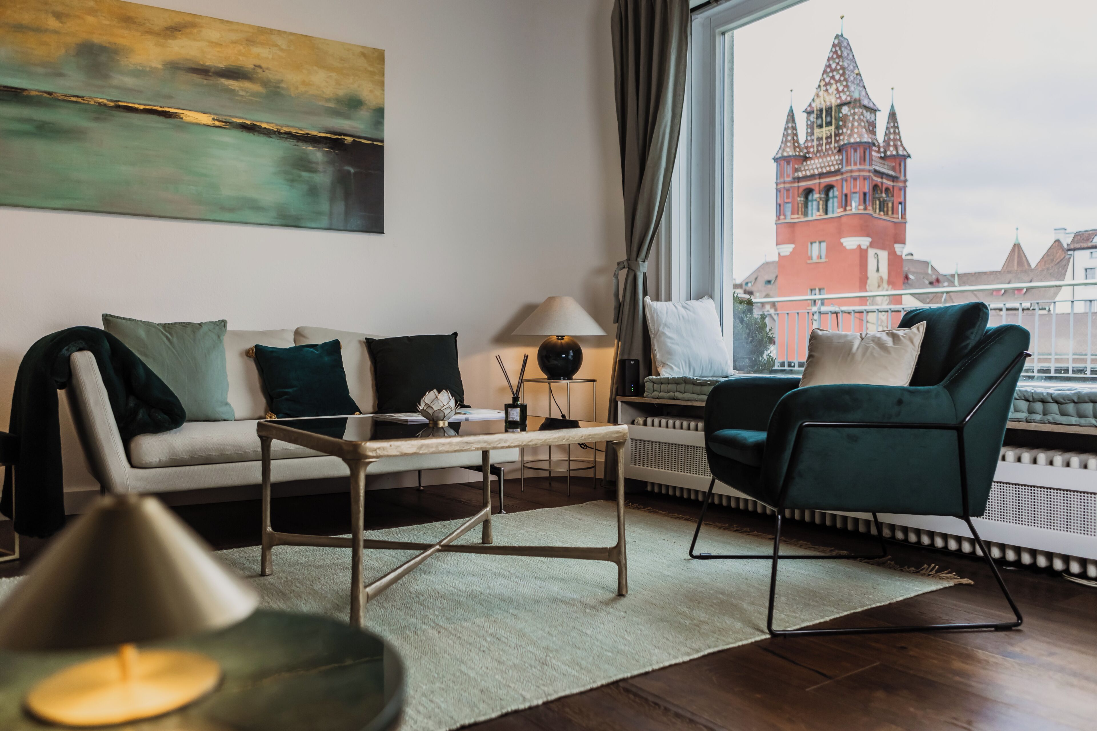 Ein modernes Wohnzimmer mit Sofa, Sessel und Couchtisch, einem Gemälde an der Wand und Blick auf ein rotes Gebäude durch ein Fenster.