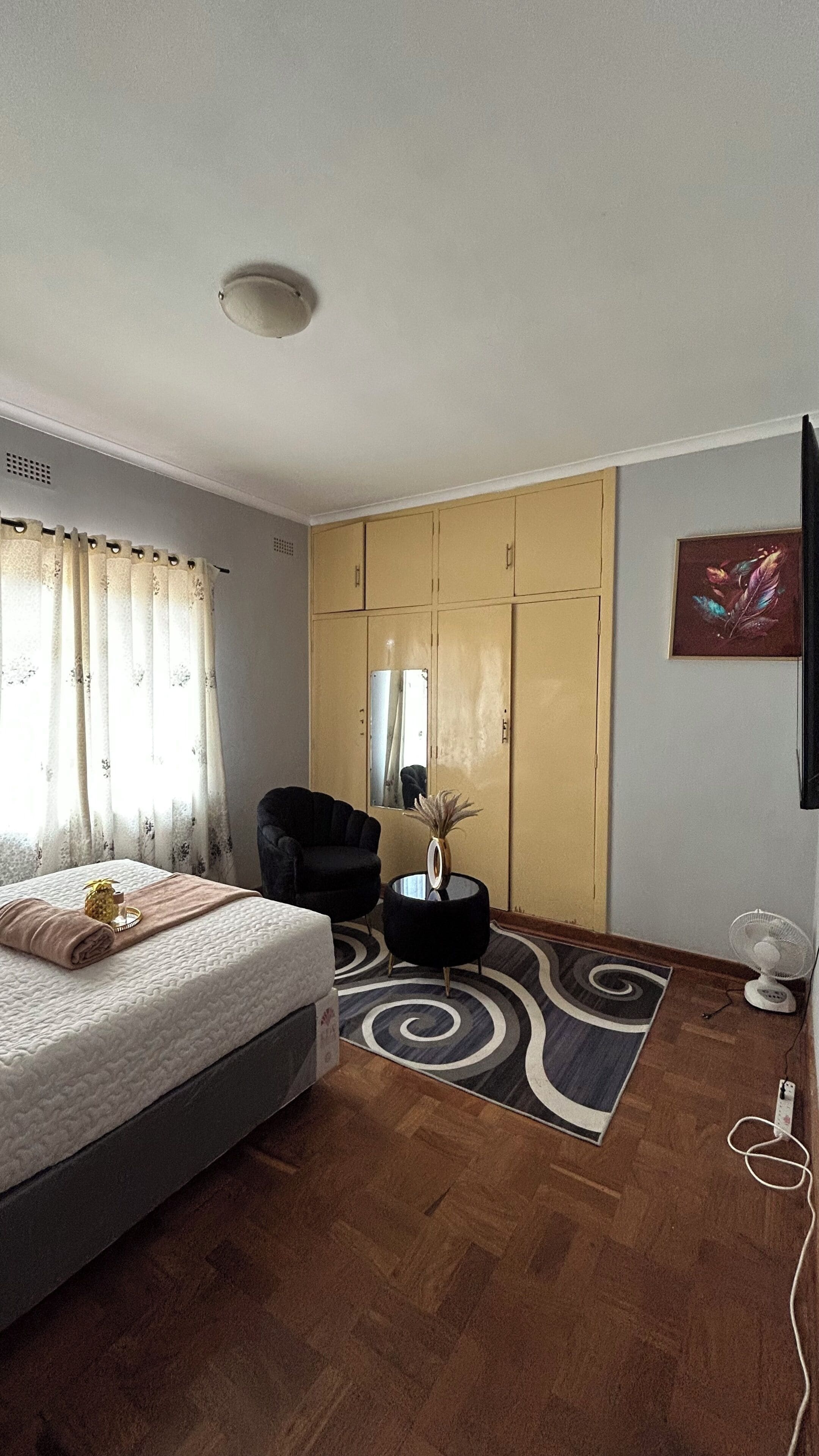 A bedroom with a bed, a chair, a wardrobe, a mirror, a fan, and a decorative vase.