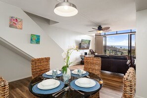 Et spiseområde med et rundt bord dækket til fire personer, omgivet af fletstole, en vase med blomster og udsigt til det frie landskab gennem store vinduer.