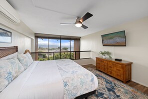 A bedroom with a large bed, a ceiling fan, a window with a view, a wooden dresser, and a painting on the wall. - Charming 2BD w Ocean Views Near Beach - KV F401 (Lahaina)