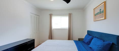 Un dormitorio con cama, ventilador de techo, armario, ventana con cortinas y un cuadro en la pared.