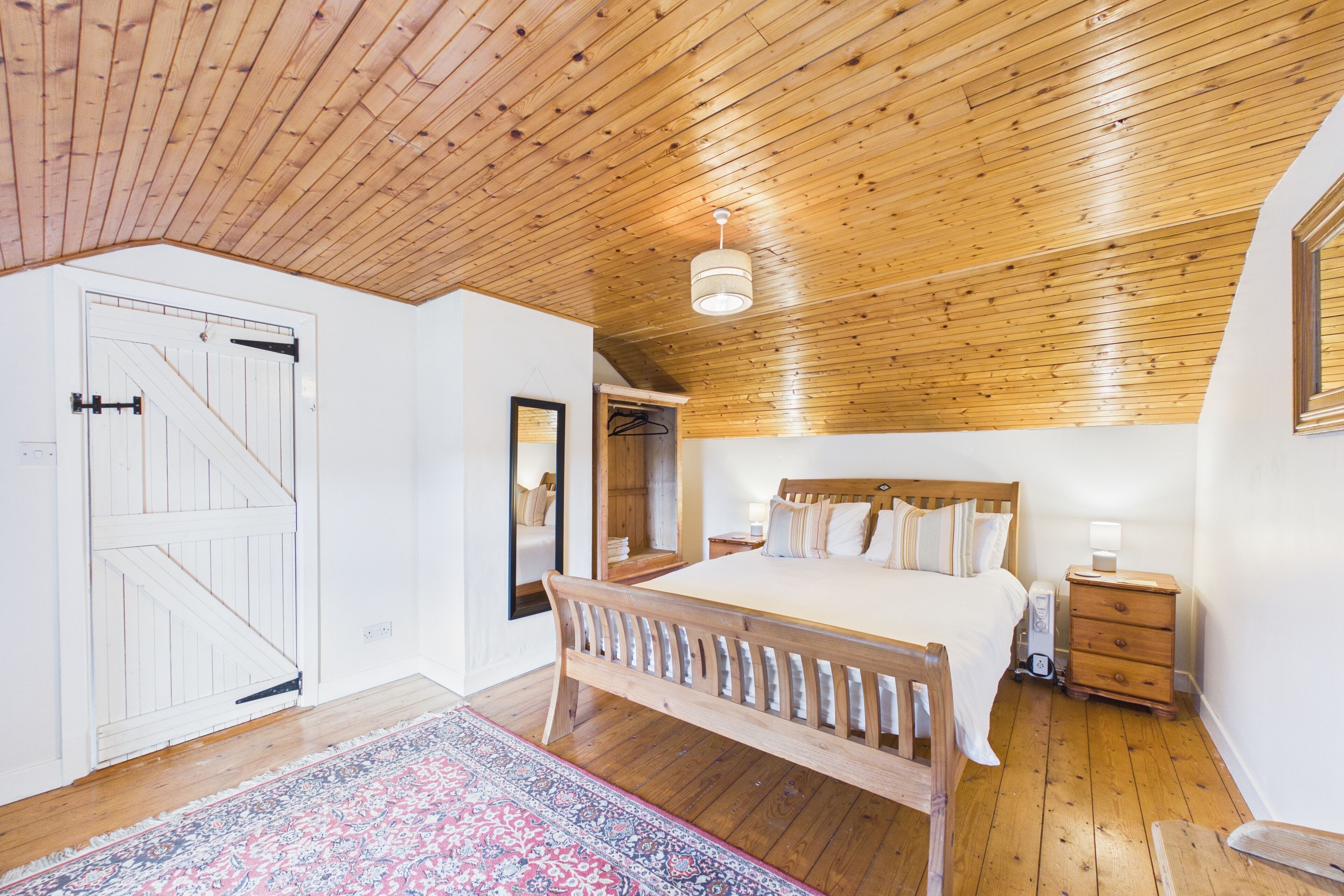 A bedroom with a wooden ceiling, a bed with a wooden headboard, a bedside table, a nightstand, and a mirror on the wall.