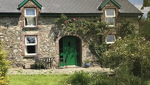 Una casa de piedra con puerta verde, un pequeño porche con sillas y un jardín delantero.
