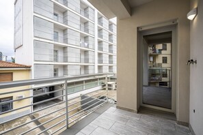 Ein Balkon mit Metallgeländer, eine Tür zu einem Innenbereich und im Hintergrund ein Gebäude mit mehreren Balkonen.