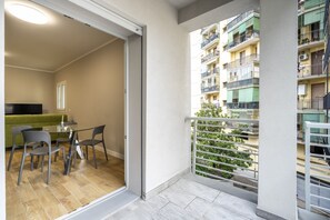En balkon med udsigt til en høj bygning, et spiseområde med bord og stole, samt en grøn sofa.