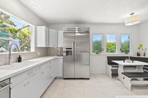 Eine moderne Küche mit Edelstahl-Kühlschrank, weißen Schränken, einer integrierten Bank und großen Fenstern.