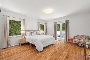 Une chambre avec un plancher en bois, un lit avec une literie blanche, une table de chevet, une chaise et une vue sur la verdure à l’extérieur.