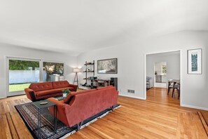 Un salon spacieux avec un canapé rouge, une table basse et une lampe. On aperçoit un coin repas à travers une porte ouverte.