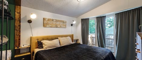 Un dormitorio con cama, dos lámparas de noche, cabecera de madera, una obra de arte en la pared y una ventana con cortinas.