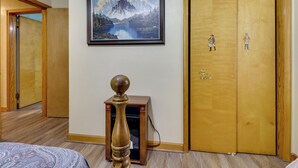A hotel room with a wooden floor, a bed with a patterned bedspread, a small wooden table with a spherical top, a framed painting of a mountain landscape, and a wooden door with a coat of arms. - Waterfront Family Home w/ Dock in Poquoson! (Poquoson)