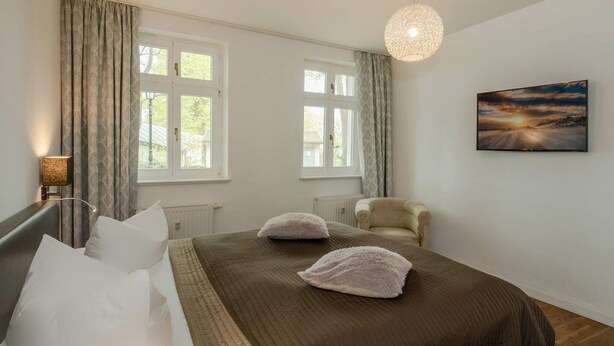 Ein Schlafzimmer mit einem Bett, zwei Fenstern, einem Sessel, einem Gemälde und einer Pendelleuchte.