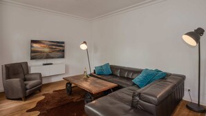 Ein modernes Wohnzimmer mit einer Ledersofa, einem Holztisch, einem Ledersessel, einer stehenden Stehlampe und einem an der Wand montierten Flachbildfernseher.