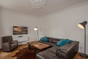 A modern living room with a leather sofa, a wooden coffee table, a leather armchair, a standing floor lamp, and a flat-screen TV mounted on the wall. - Villa Hohe Buchen, Apartment 02 with a garden, just 150 m from the beach - Beautiful (Heringsdorf)