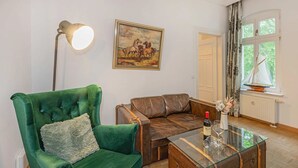 Um quarto com uma poltrona verde, um sofá de couro marrom, uma mesa de centro de vidro com uma garrafa e copos, uma pintura de cavalos e uma janela com cortinas.