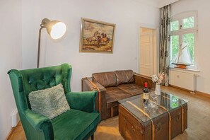 A room with a green armchair, a brown leather sofa, a glass coffee table with a bottle and glasses, a painting of horses, and a window with curtains. - Villa Hohe Buchen, Apartment 03, just 150 meters from the beach—A beautiful vacation rental (Heringsdorf)