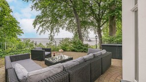 En udendørs terrasse med fletningsmøbler, et glasbord og havudsigt.